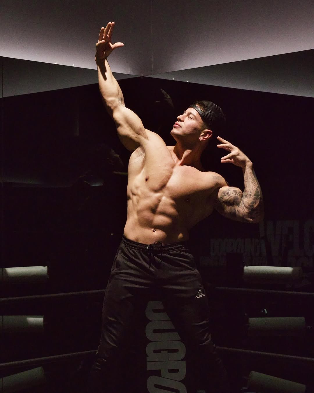 Shirtless man in a gym striking a dynamic pose with one arm raised and the other bent, wearing black pants and a backwards cap.
