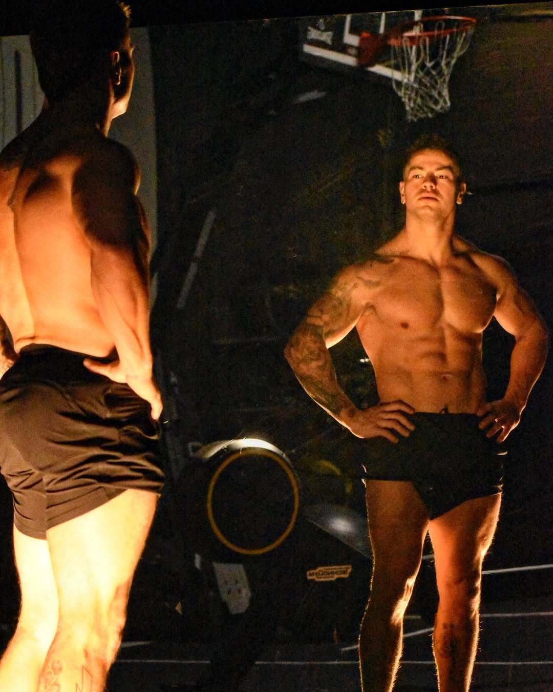 Shirtless muscular man looking at his reflection in the mirror inside a dimly lit gym, with a basketball hoop in the background.