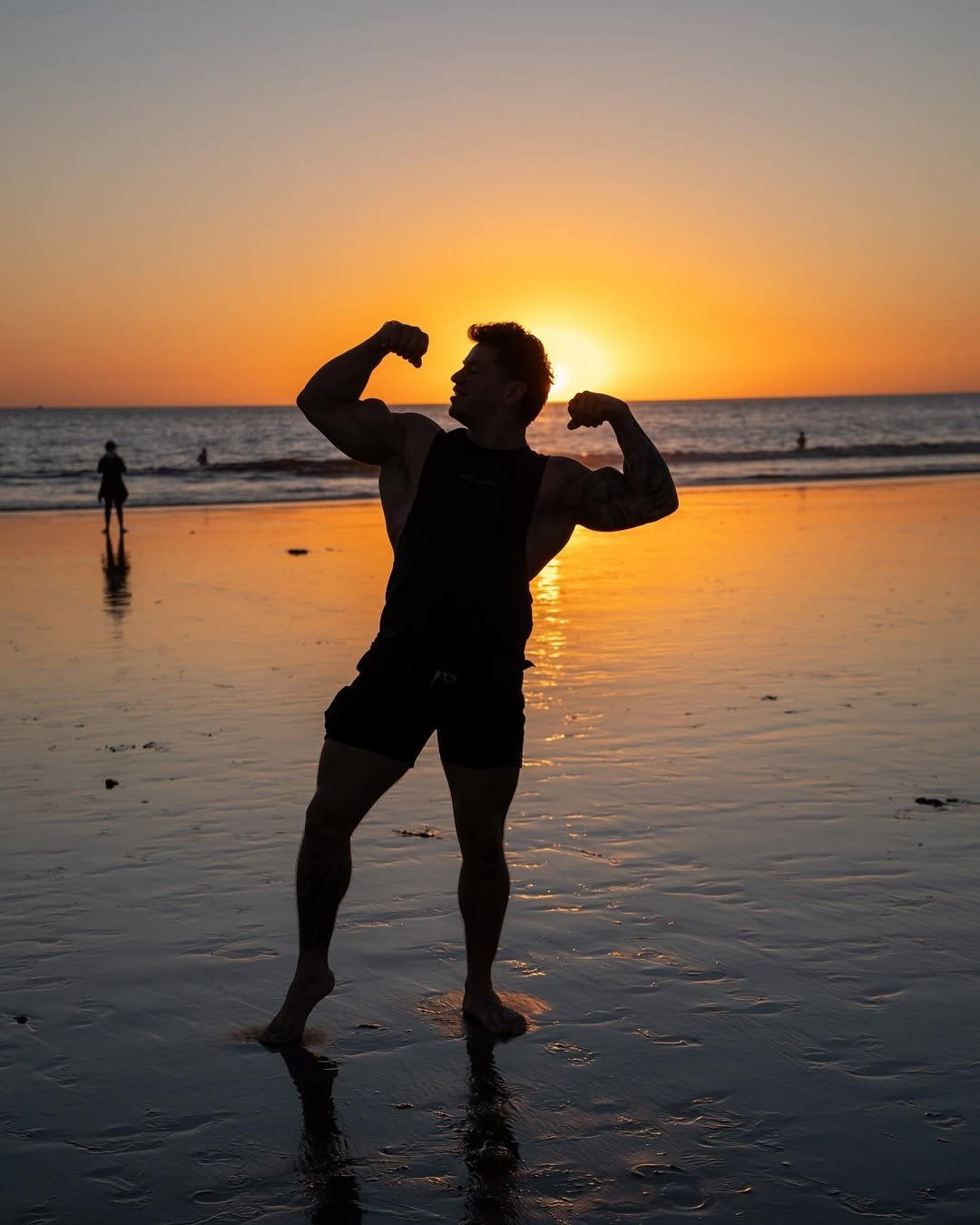 Cristian Castaño flexing both arms at sunset on a beach, silhouetted against the orange and purple sky.