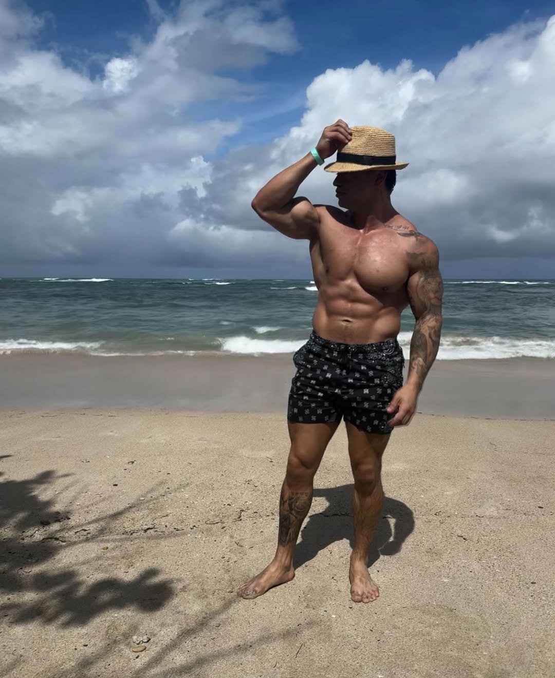 Cristian Castaño standing on the beach, adjusting a straw hat with both hands, showing his defined physique under a cloudy sky.