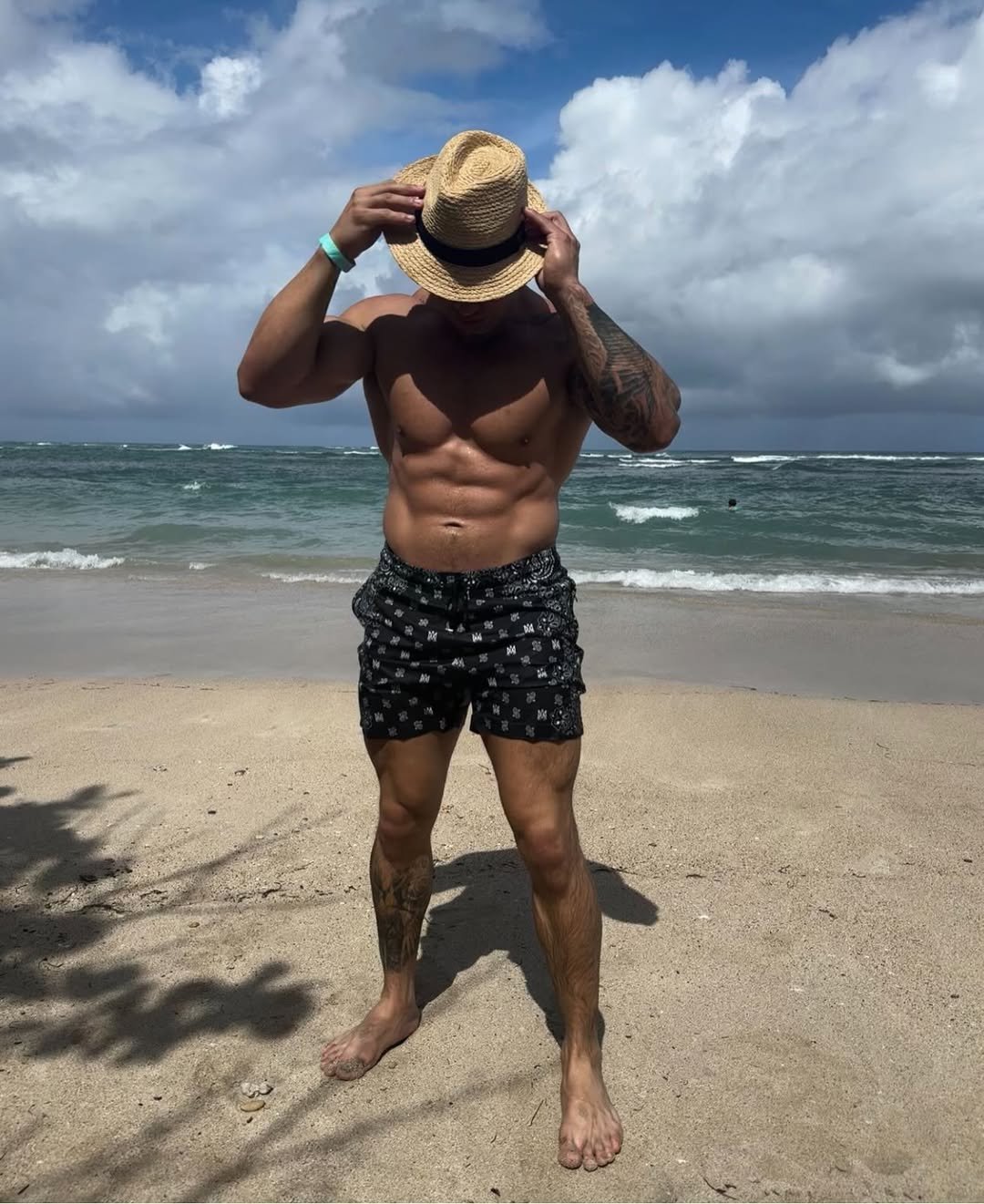 Cristian Castaño posing on the beach with a straw hat, flexing slightly and looking toward the ocean.