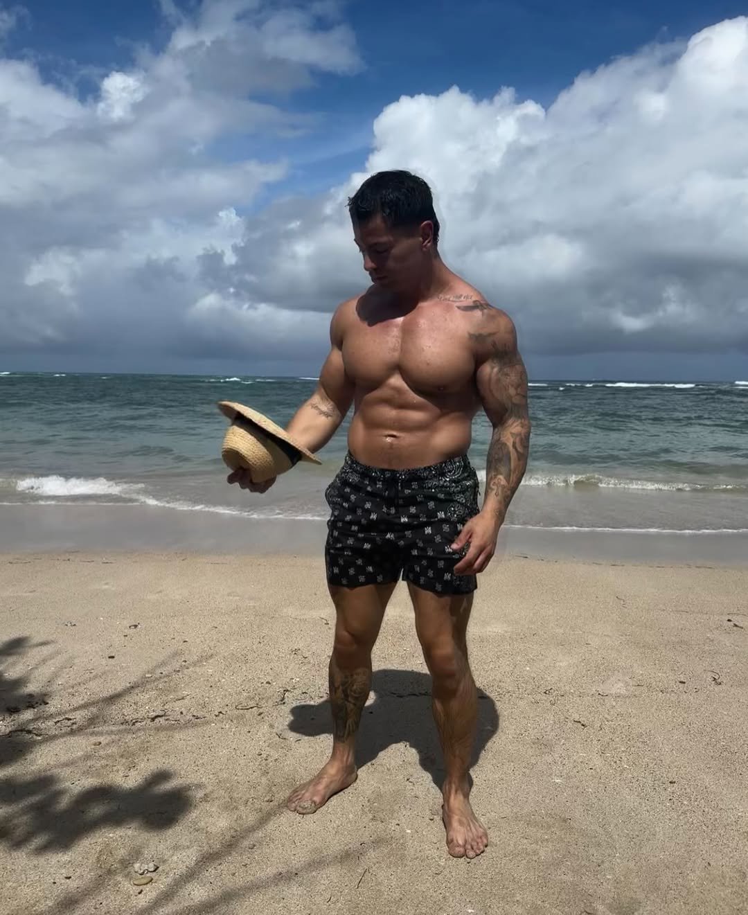 Cristian Castaño looking down at a straw hat in his hand while standing shirtless on a beach, waves in the background.