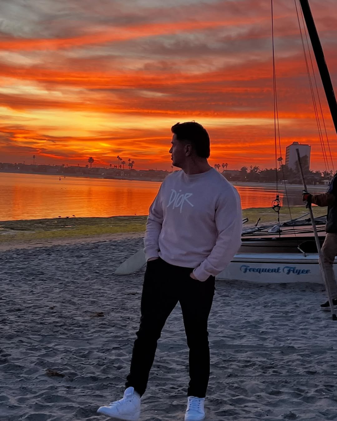 Cristian Castaño standing on a beach at sunset, wearing a light Dior sweatshirt and white sneakers, looking toward the fiery orange sky.