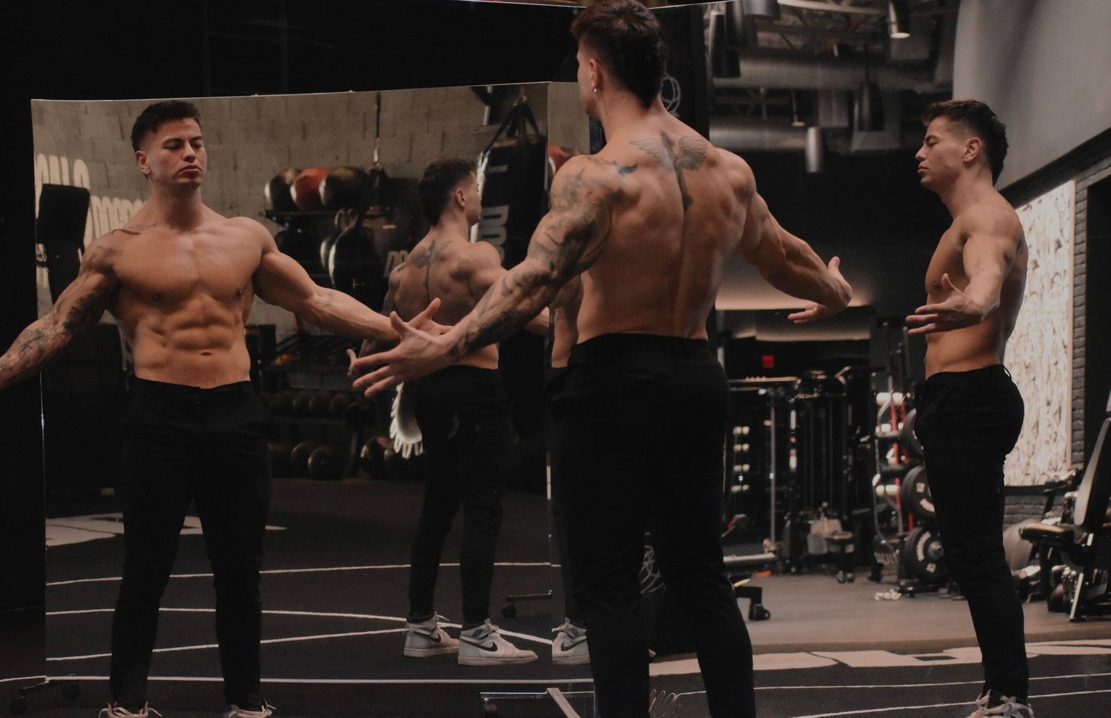Christian Castano, a muscular male fitness influencer, posing shirtless in a gym with his arms outstretched to the sides, reflected in a large mirror.