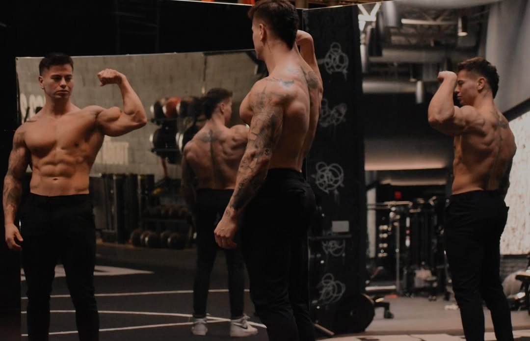 Cristian Castaño shirtless in the gym, flexing his arm while looking at his reflection in a mirror, surrounded by a fitness environment.