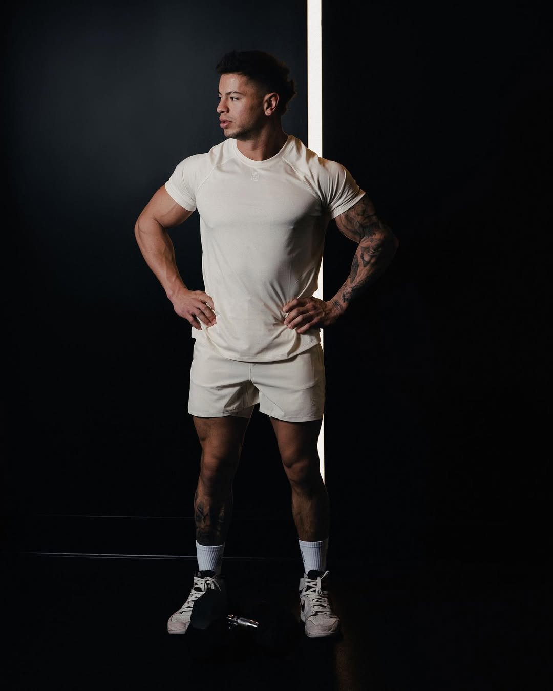 Cristian Castaño standing with hands on hips in a cream-colored gym outfit, in front of a black background with a vertical light beam.