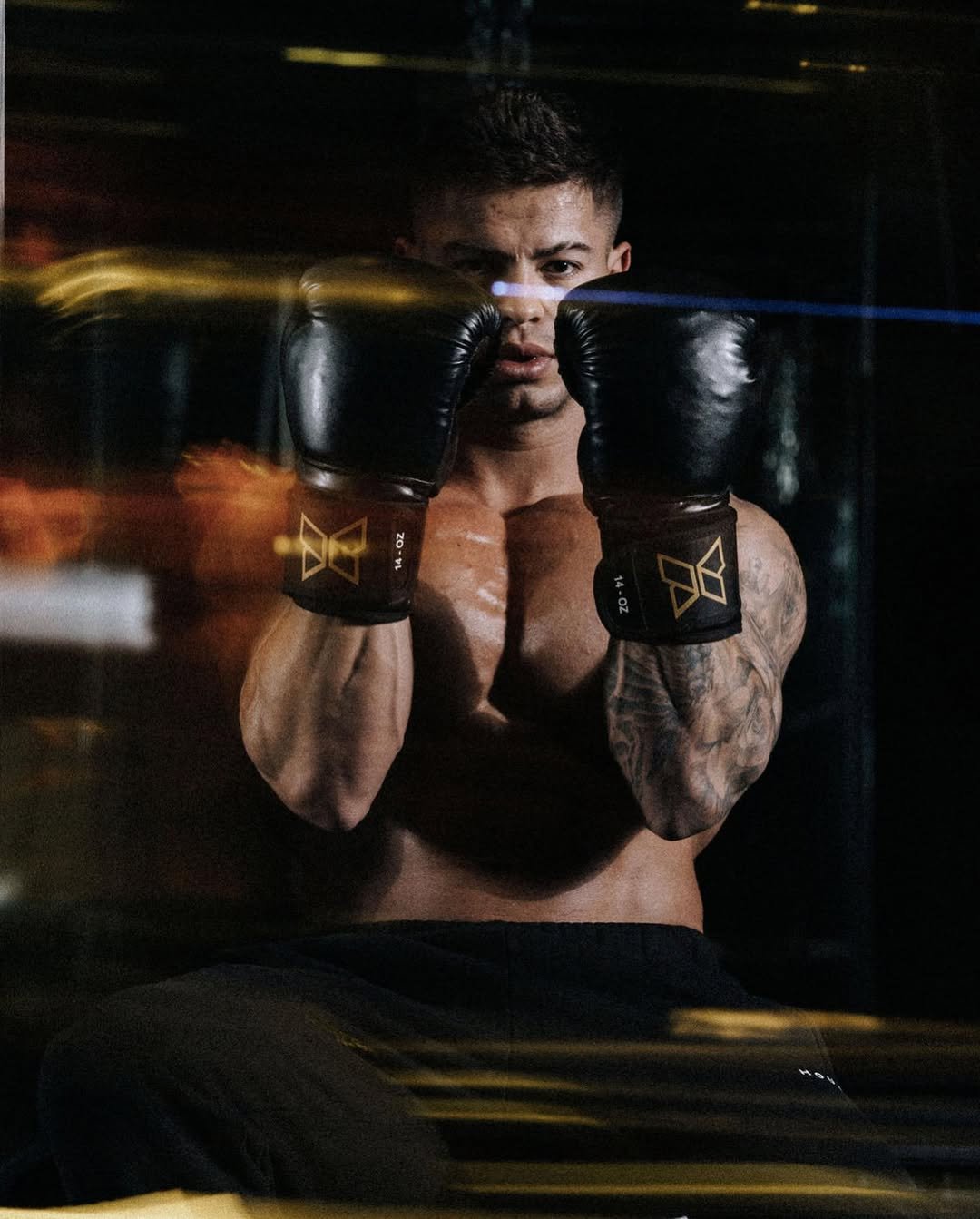 Cristian Castaño shirtless with black boxing gloves raised in front of his face, captured through a dynamic motion blur effect.