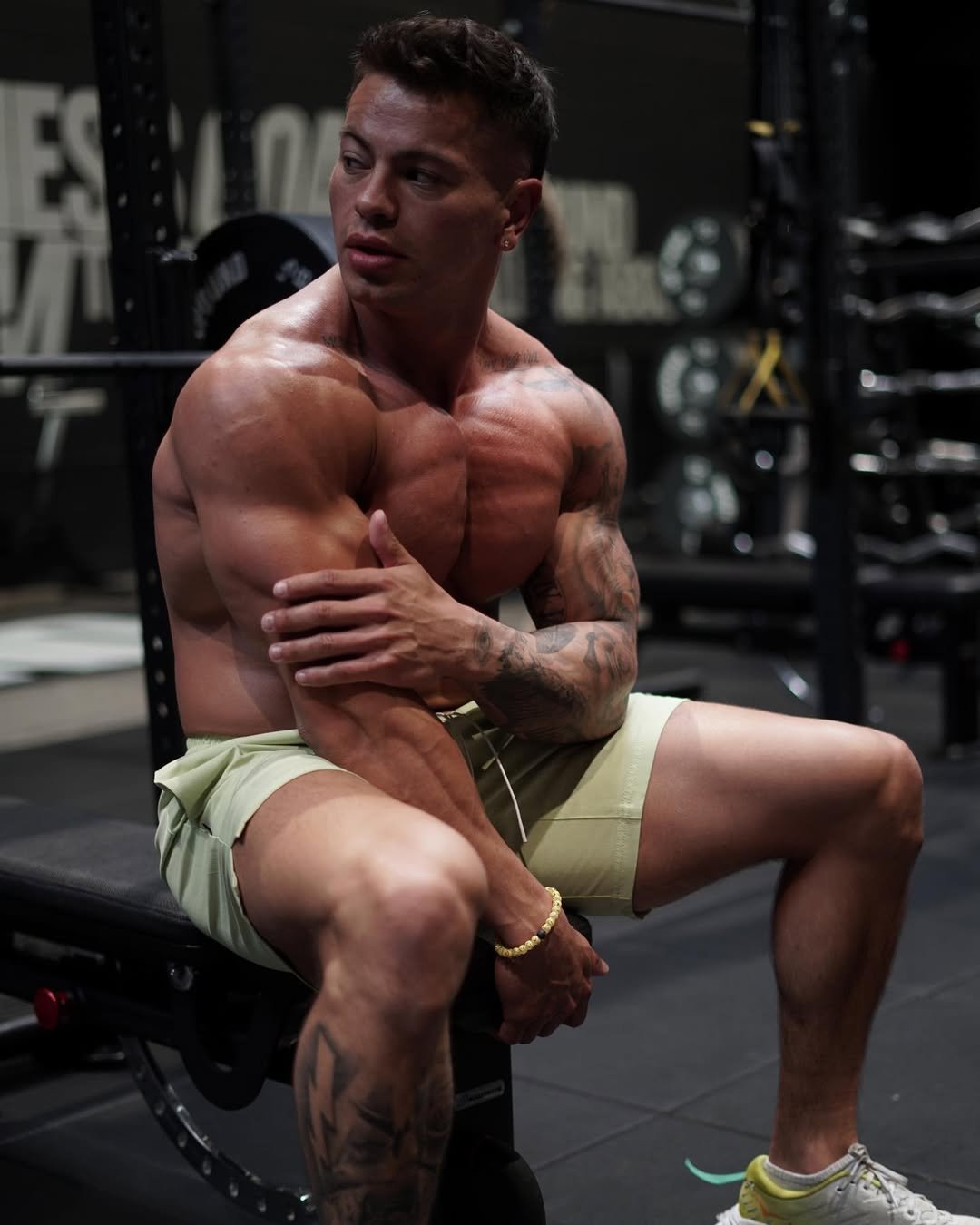 Cristian Castaño resting on a gym bench, shirtless and touching his arm, showing muscle definition and focus.