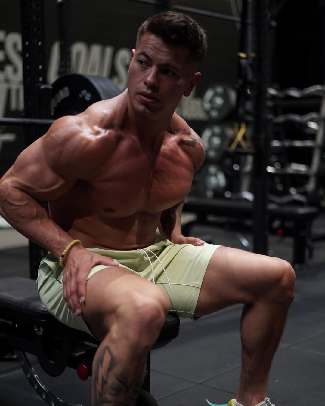 Cristian Castaño sitting shirtless on a gym bench, looking to the side with a focused expression, wearing light green shorts.