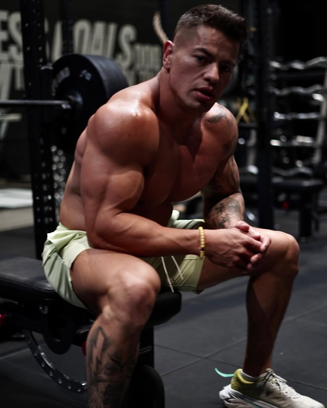 Cristian Castaño looking directly at the camera while seated shirtless on a gym bench, hands clasped and resting on his legs.