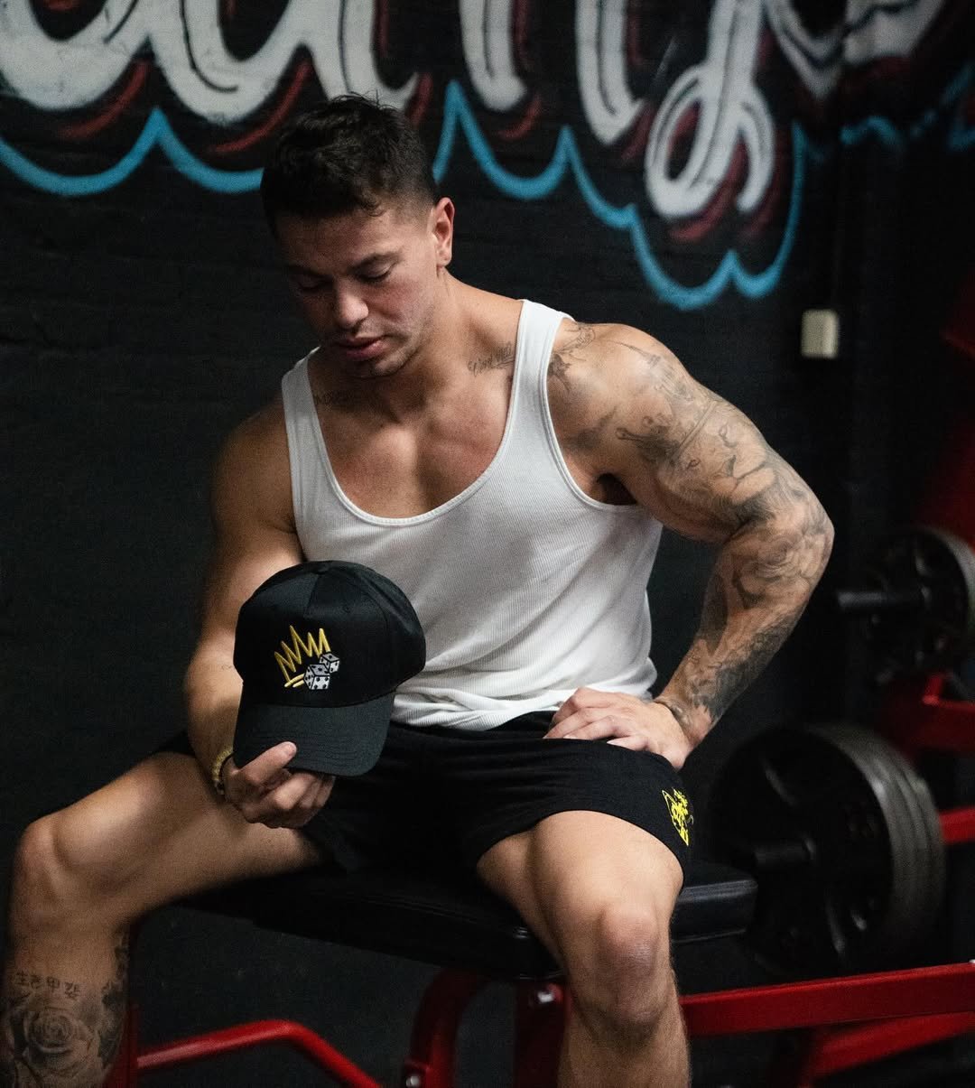 Cristian Castaño sitting on a gym bench, wearing a white tank top and black shorts, holding a black cap with a gold crown logo.