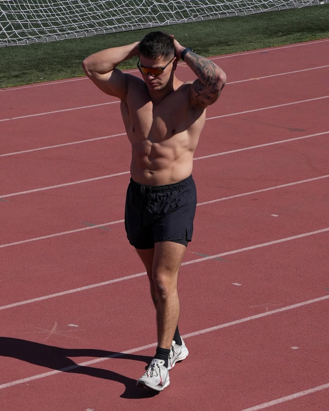 Cristian Castaño walking shirtless on a track field with his hands on his head, wearing black shorts, sunglasses, and white running shoes.